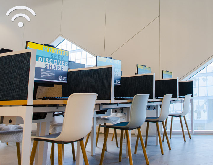 background image: A table with computers and privacy dividers. There are chairs available for each computer.