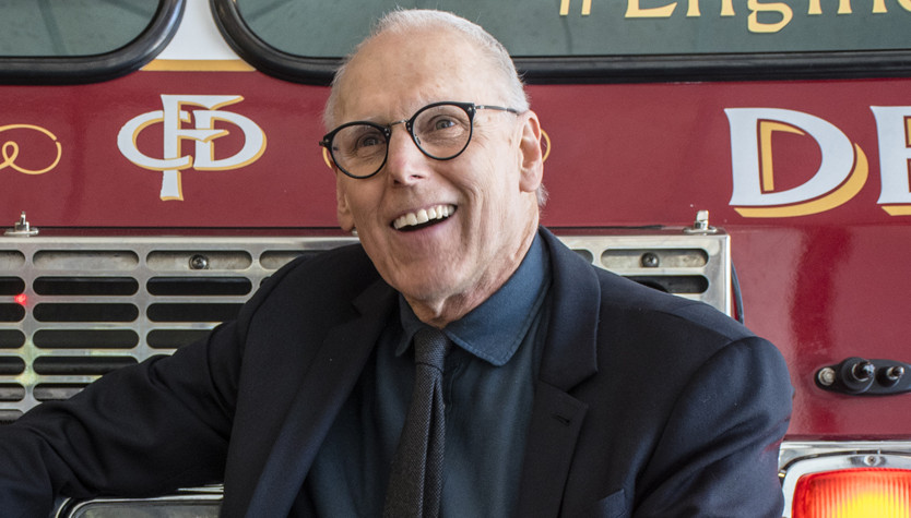 Photograph of Bill Ptacek sitting in front of a fire engine. 