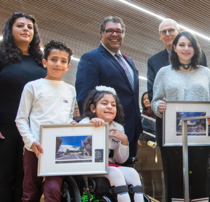 A group of people of various ages standing on a large staircase each holding a framed photograph. 