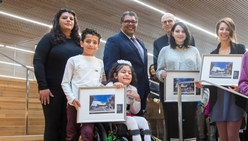 A group of people of various ages standing on a large staircase each holding a framed photograph. 