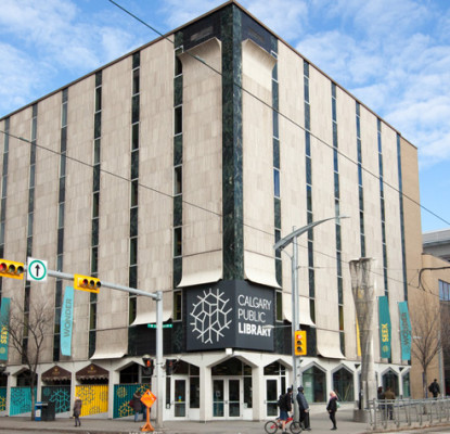 Photograph of a building from the street with a light beige exterior and a large grey sign that reads "Calgary Public Library."