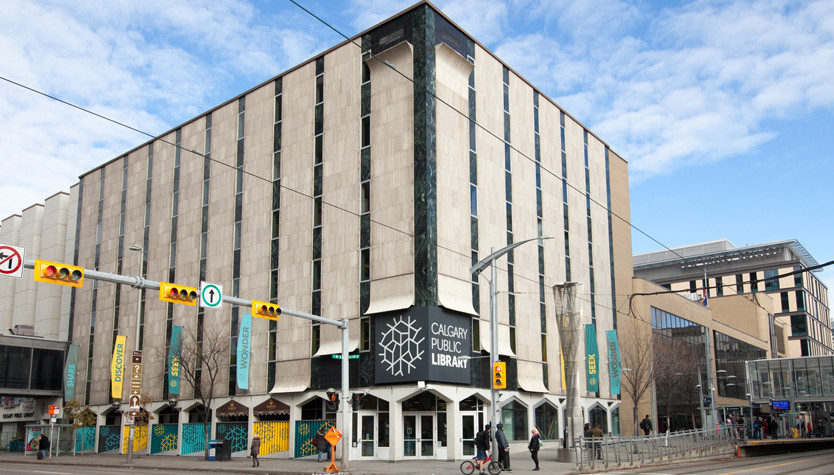 Photograph of a building from the street with a light beige exterior and a large grey sign that reads "Calgary Public Library."