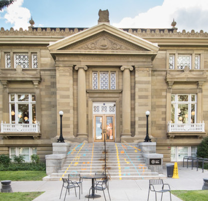 A two story sandstone building with large pine trees.