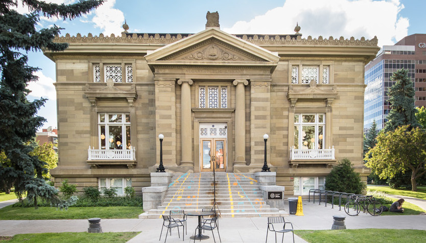 A two story sandstone building with large pine trees.