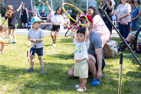 Children and adults participate in a hoop dance at a Powwow. A young child in the foreground holds up a colorful hoop with determination, while other children dance with their hoops in the background. Adults and spectators enjoy the lively atmosphere on a sunny day.