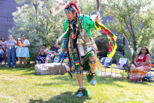 An Indigenous dancer in traditional regalia performs at a Powwow, showcasing vibrant green attire adorned with intricate beadwork and feathers. The dancer is in mid-step, holding a staff and wearing moccasins with fur and jingles. Spectators in colourful clothing watch and capture the moment on their phones, sitting and standing in a grassy outdoor area.