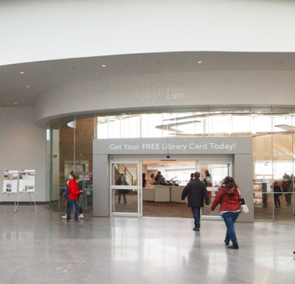 A large open modern space with a set of doors leading into a library. Several people are walking towards or entering the library.