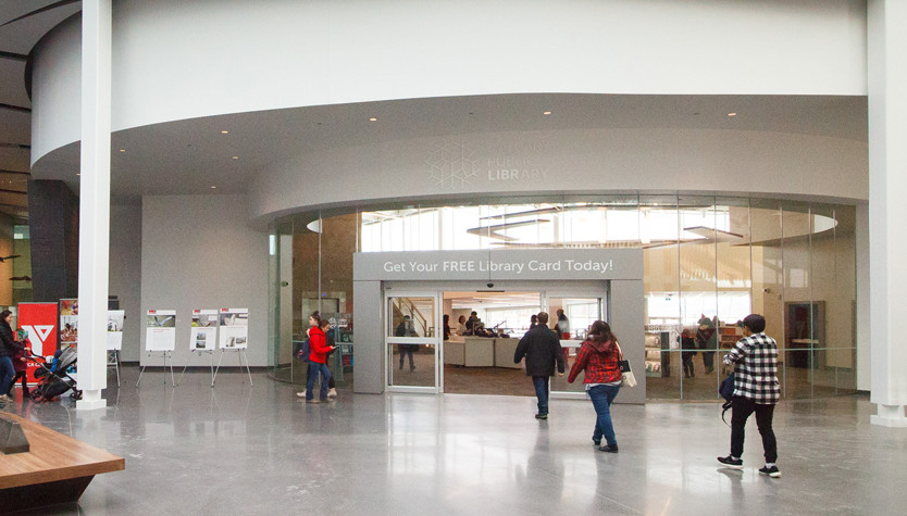 A large open modern space with a set of doors leading into a library. Several people are walking towards or entering the library.