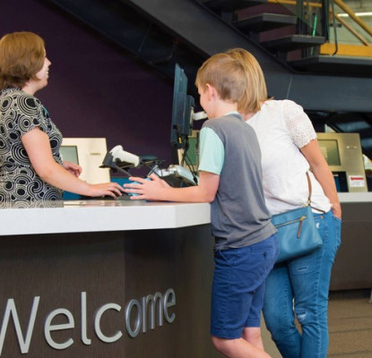 Photograph of the interior of a Library location. Three people stand in line at the counter while a staff member helps them. 