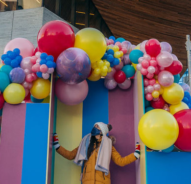 A person standing under an arch of colourful balloons in an urban setting.