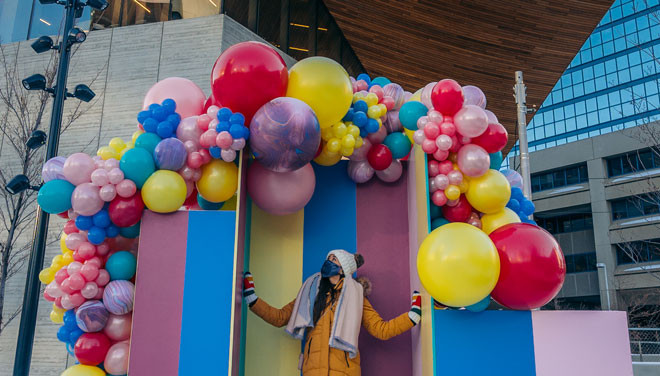 A person standing under an arch of colourful balloons in an urban setting.