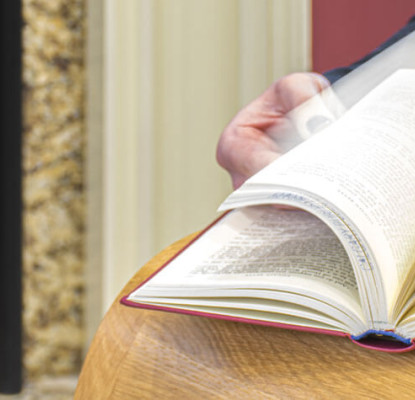 Photograph of a person flipping through a book on a wooden table. They are sitting near a fireplace. 
