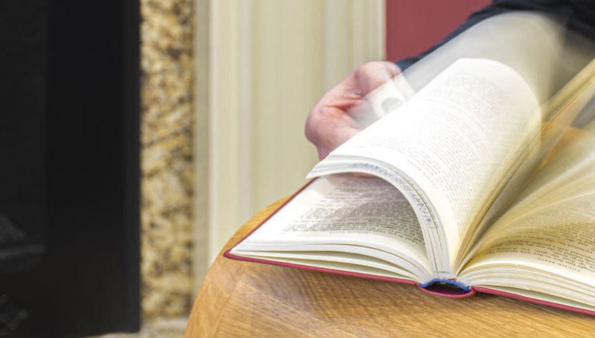 Photograph of a person flipping through a book on a wooden table. They are sitting near a fireplace. 