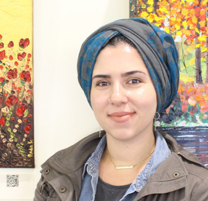 Nada Khatib standing in front of two paintings. One is of red flowers and the other is trees and water.