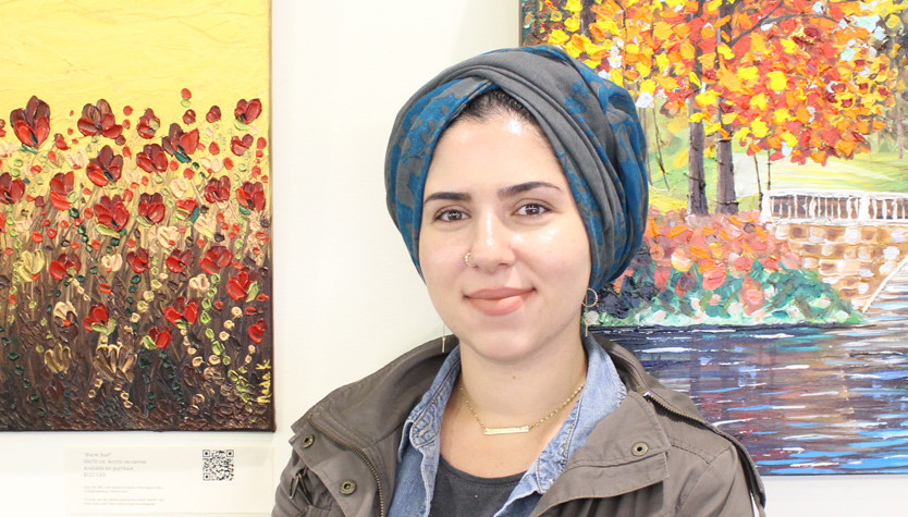 Nada Khatib standing in front of two paintings. One is of red flowers and the other is trees and water.