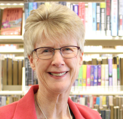 Photograph of Marje Wing standing in front of a bookshelf. 