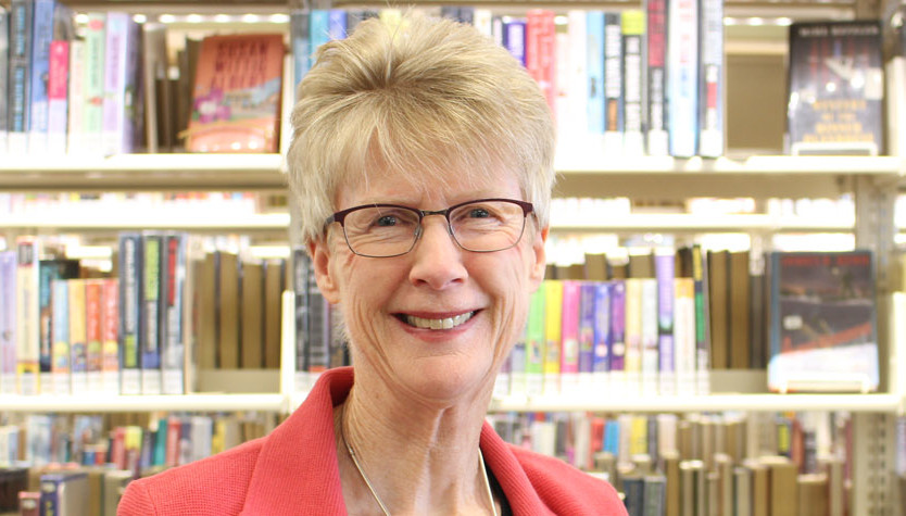 Photograph of Marje Wing standing in front of a bookshelf. 