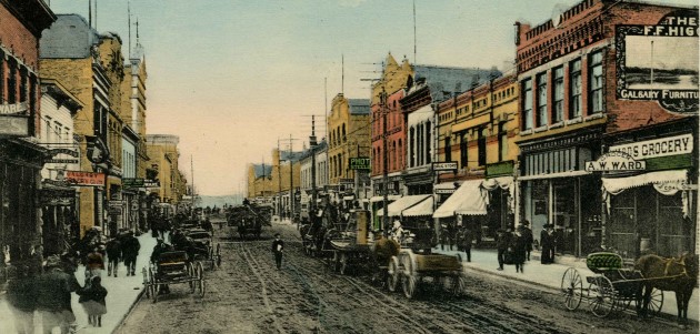An old photograph of a city street. There are people walking and horses and carriages on the street.