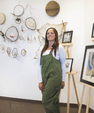 A photograph of a woman in green overalls standing in front of her artwork on easels. 