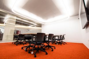 Photograph of a meeting room. There is an orange carpet, and a couple of long tables surrounding them are chairs.