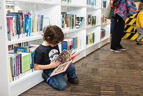 A child is sitting on the floor in front of a bookshelf, flipping through a comic book.