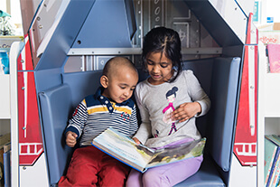 A toddler and a child are sitting in a reading nook, flicking through a picture book.