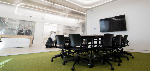 A photograph of a meeting room. In the centre of the room is a table with chairs. In the background is a tv mounted on the wall.