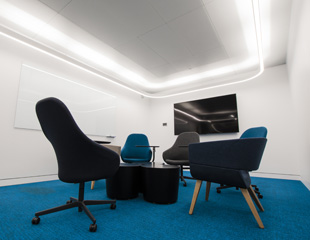 A photograph of a meeting room. Rolling chairs surround a black table, and a TV is on the wall in the background.