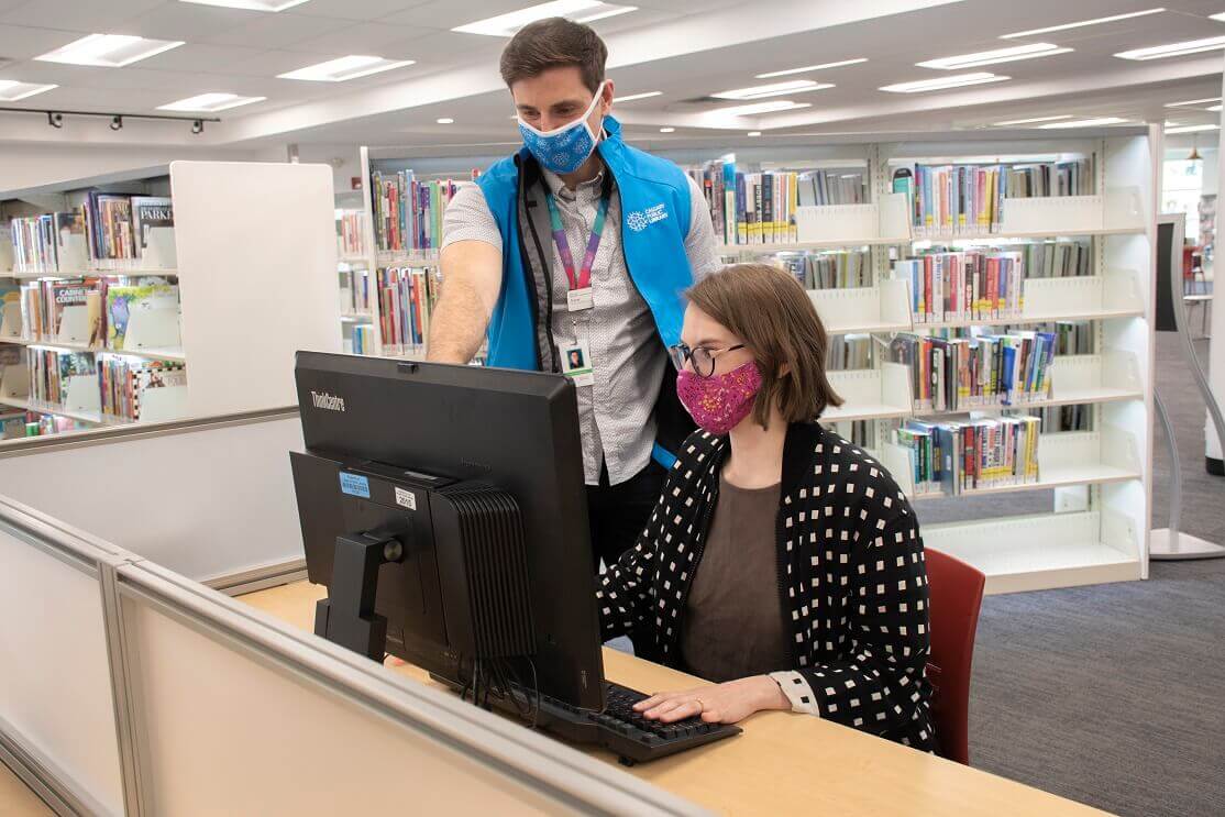 background image: Photo of two people working at a computer. One person is standing, the other is sitting. 