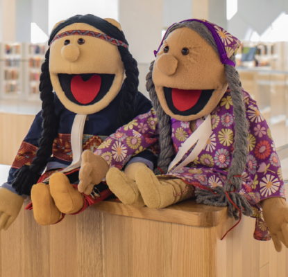 An image of two puppets designed as indigenous people with an overlay of a graphic of a table with math work sheets and a cup with pencils.