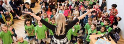 A woman with her back to the camera has her arms spread out in the air in front of a large group of children.