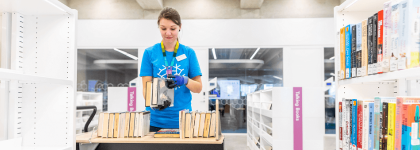 A Library assistant, wearing a blue shirt and black gloves, is organizing books on a cart in a modern library. She is holding a stack of books while arranging others that are lined up on the cart. The shelves around her are mostly empty, but some are fill