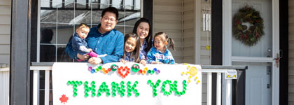 A family of five standing behind a porch holding a big banner that says Thank you on it.