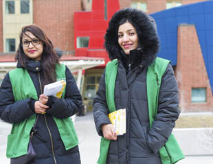 Two women in winter coats and green vests stand outside a colourful building.
