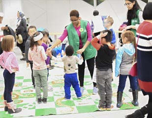Two women in green vests help a group of children play an oversized boardgame.