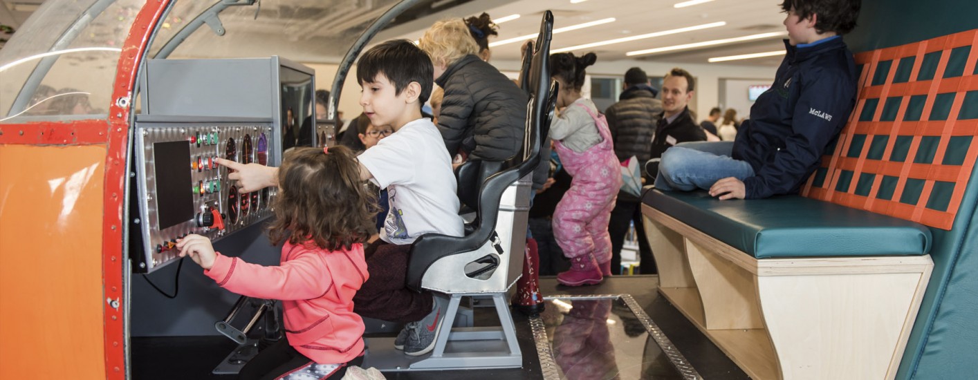 An interactive helicopter. Inside it are four different children. A little girl in the front is wearing a pink hoodie and is flicking a switch. Beside her is a young boy in a white shirt pushing a button on the console. 
