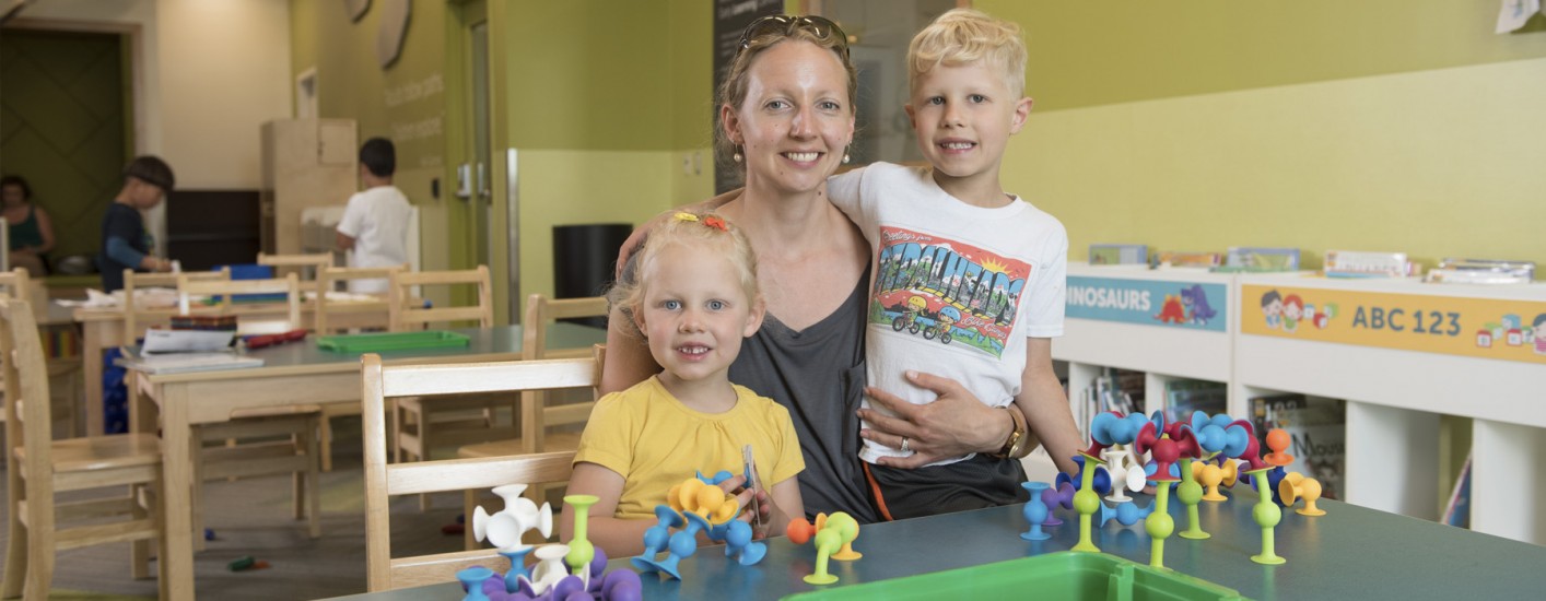 A mom and her son and daughter are sitting at a table. In front of them are a selection of toys that they are playing with.