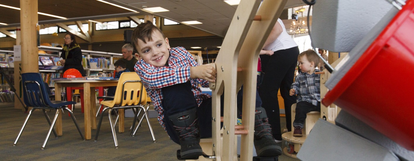 A child is holding onto a piece of equipment and is smiling. The equipment has a bucket on the end of it, and inside the buckets are gray-looking blocks that the child is moving.