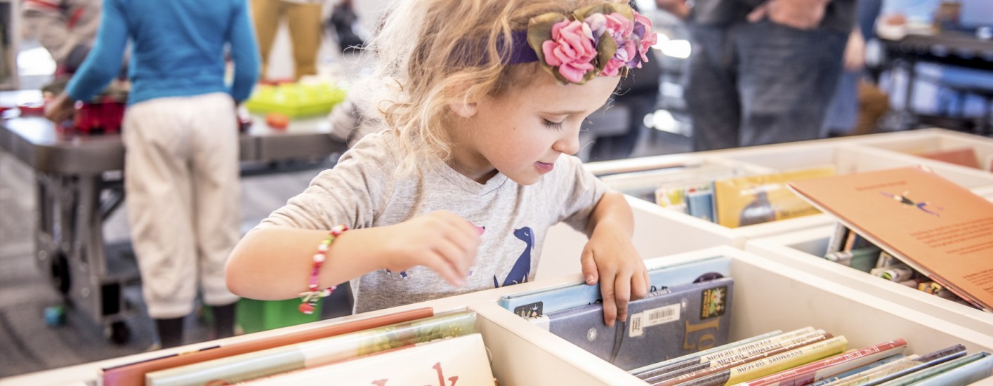 A little girl in a gray shirt, wearing a beaded bracelet and a flower crown on top of her head, is looking through a bin of picture books.