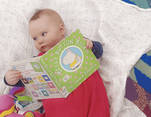 A baby holding a picture book, lying on a blanket and facing away from the camera. 