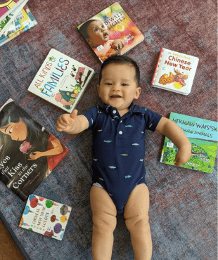 A toddler lying on a grey and red blanket. Picture books surround the toddler, and he is reaching toward the camera with his right hand. 
