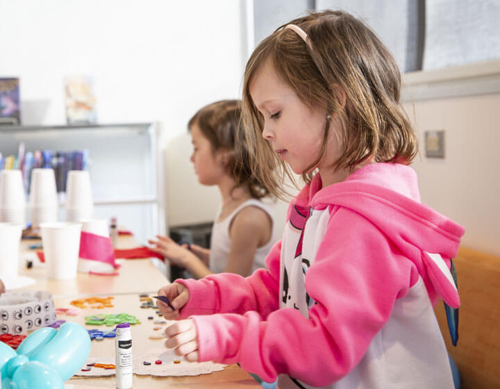 background image: A little girl in a pink and white hoodie is making a craft. She is holding a piece of construction paper and has a glue stick beside her. On her other side is another little girl cutting a piece of paper in a white tank top. 