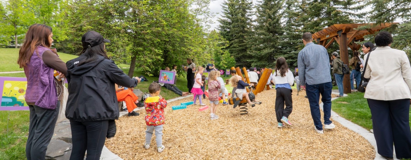 Children and adults gather in a wooded outdoor play area with wooden climbing structures, spring riders, and colourful activity signs.