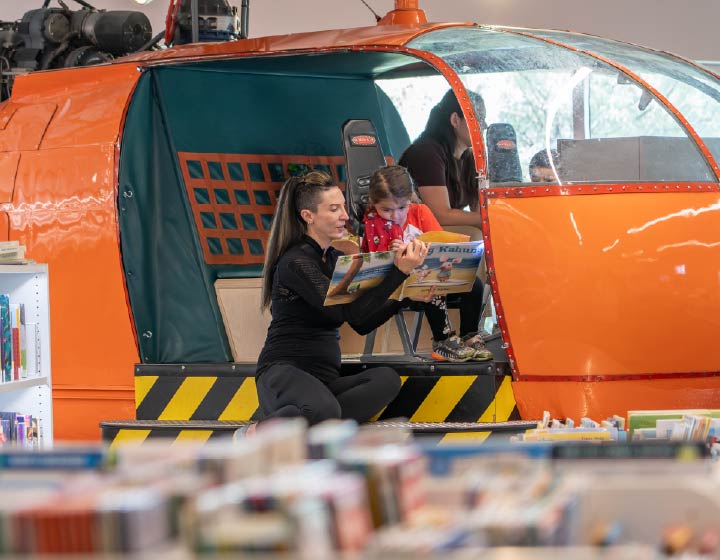 background image: Adult reading a picture book with a child inside a library play structure.
