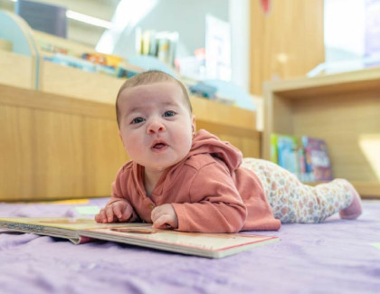 Baby lying on a blanket and looking at an open board book in a library.