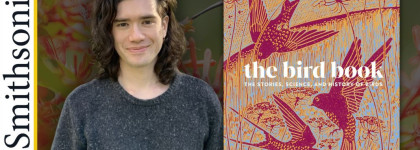 A man in a grey sweater stands beside an image of a book titled The Bird Book