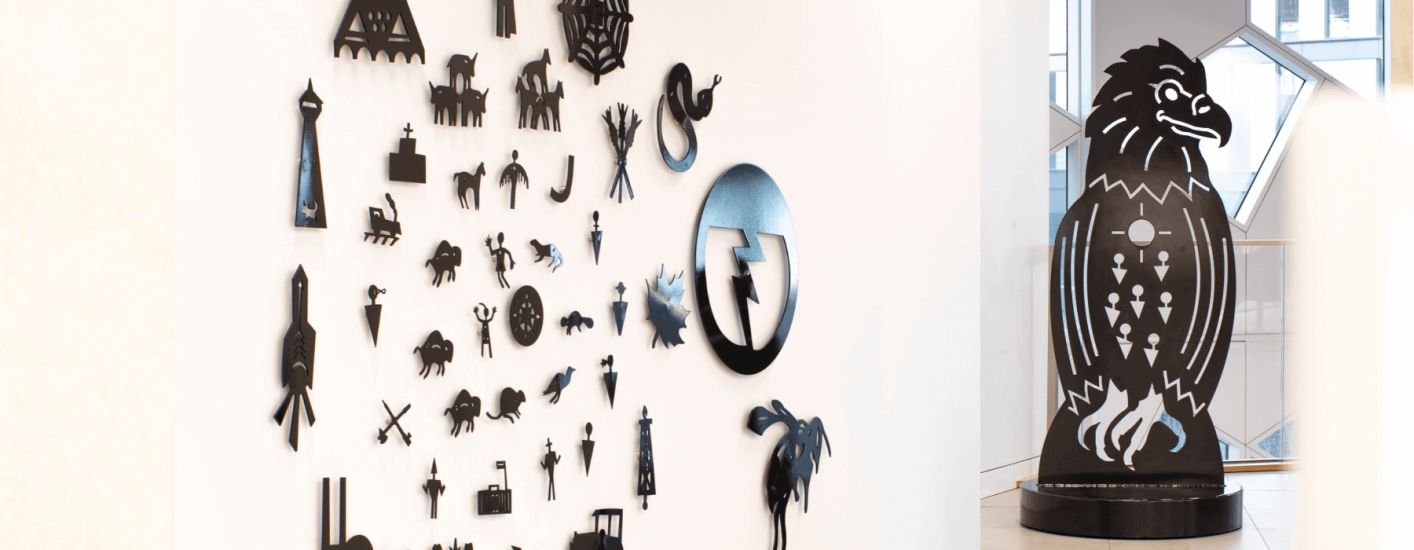 A stylized black eagle sculpture stands in front of a wall display of black symbols. The eagle faces right with its wings folded and features geometric patterns on its chest.