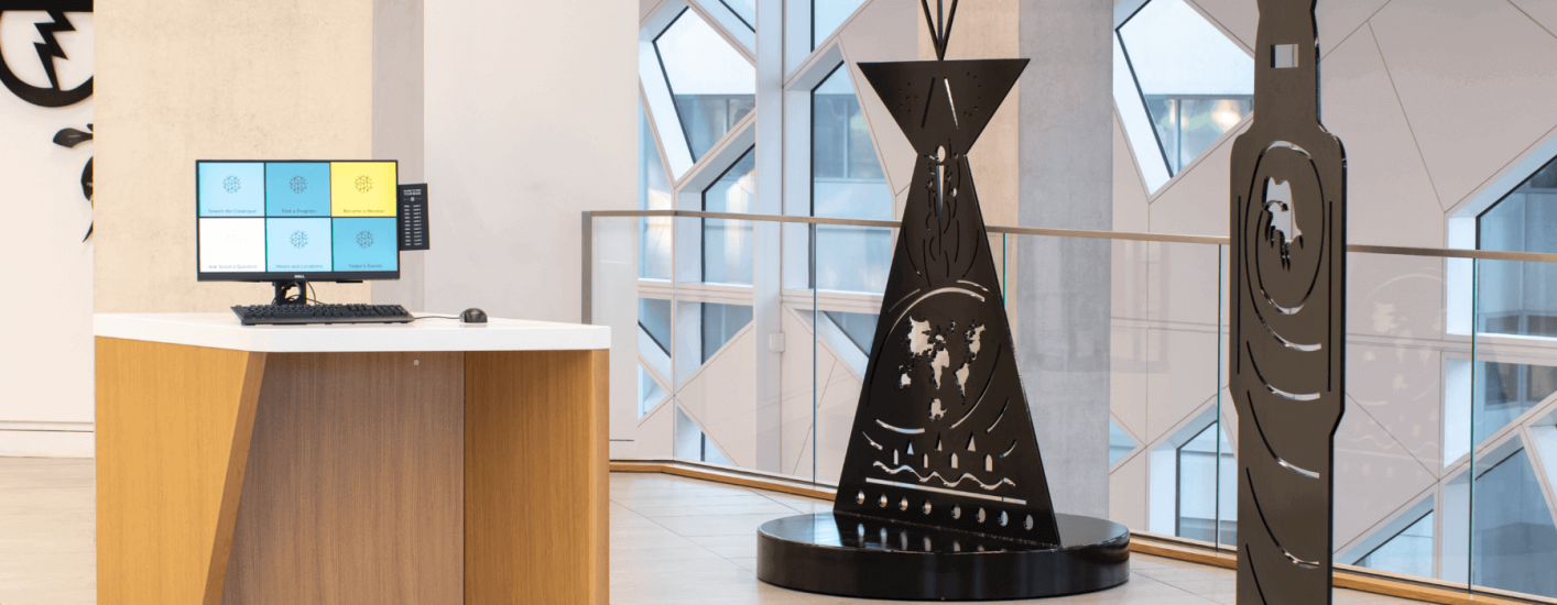 A stylized black teepee sculpture, adorned with a world map and wavy lines, is positioned in a modern building. A computer monitor sits on a nearby counter.