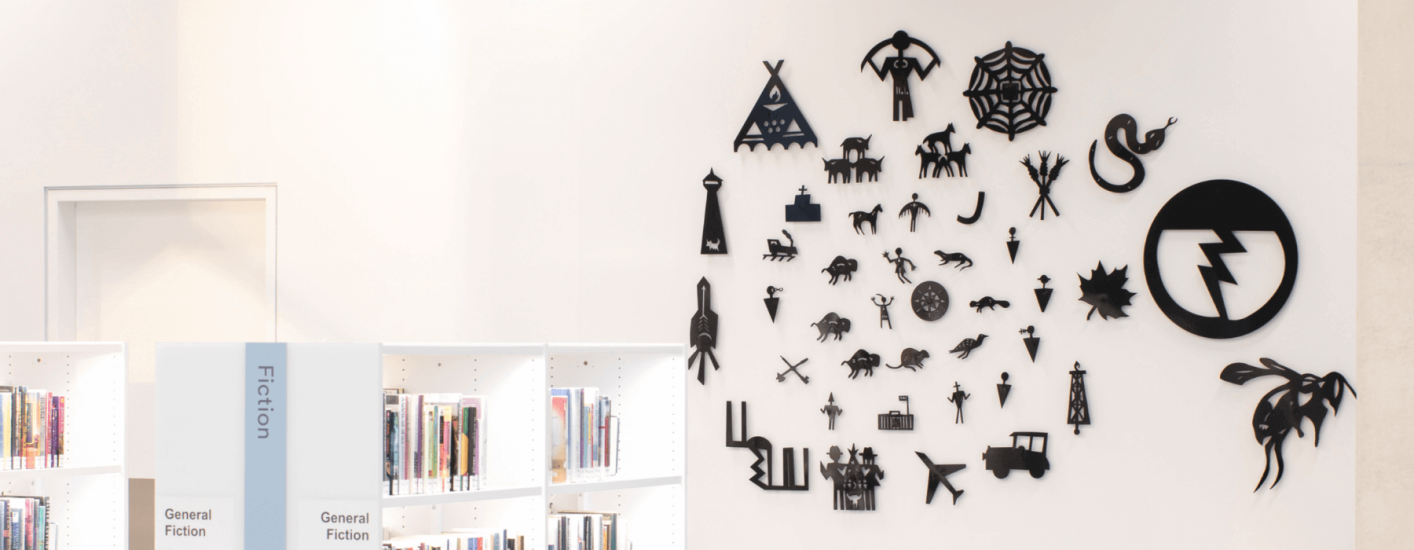 A collection of black symbols, representing various aspects of First Nations culture, decorates a white wall. Below, shelves filled with books are partially visible, with the label Fiction indicating the section.