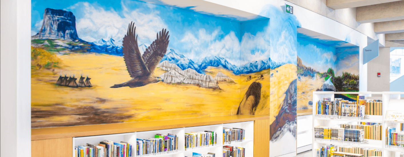 A vibrant mural spans the wall above Library bookshelves, depicting a majestic eagle soaring over a sweeping landscape of mountains and plains. The colourful scene evokes a sense of natural beauty and rich history of the region.